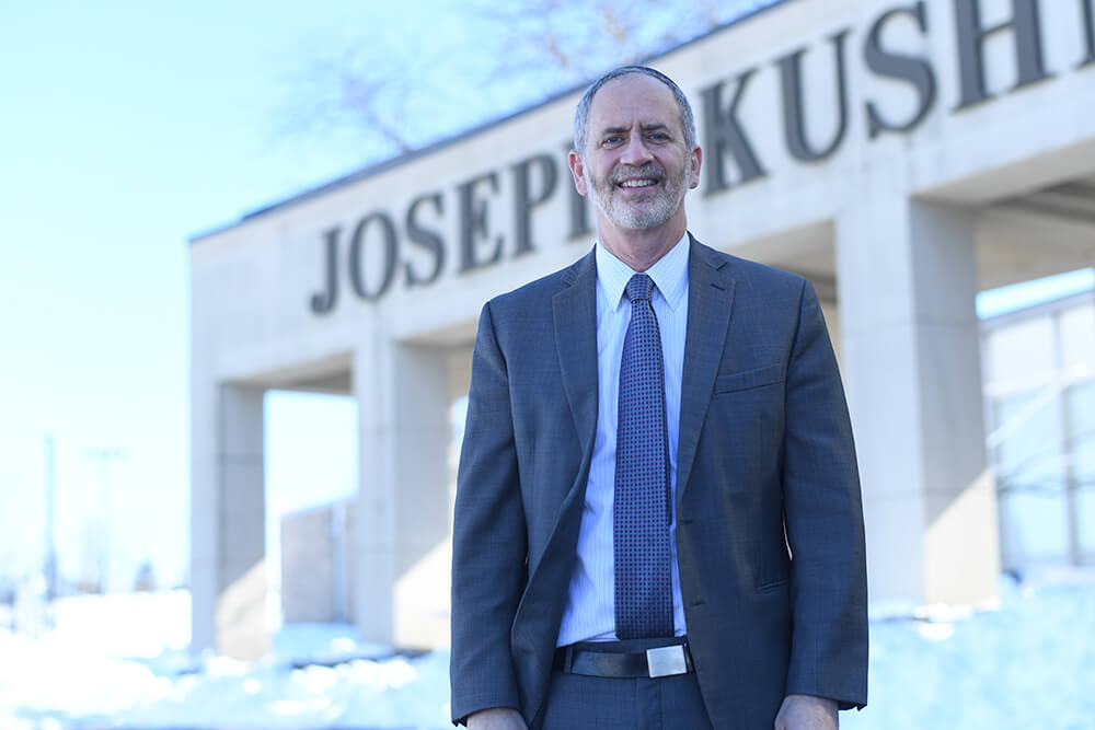 Rabbi Eliezer Rubin standing in front of Kushner Academy with the words Joseph Kush(ner) on the building behind him.